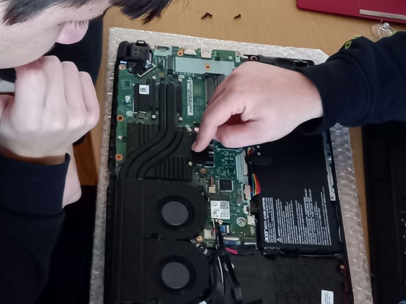 Zwei Schüler arbeiten an einem zerlegten Laptop. Die Rückseite des Laptops ist geöffnet, man sieht Bauteile